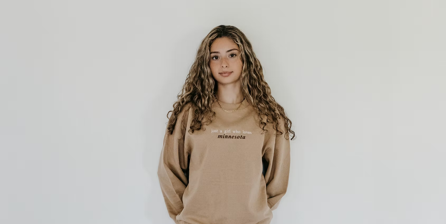 Woman wearing a beige sweatshirt with text on front stating Just a girl who loves Minnesota, on a plain backdrop